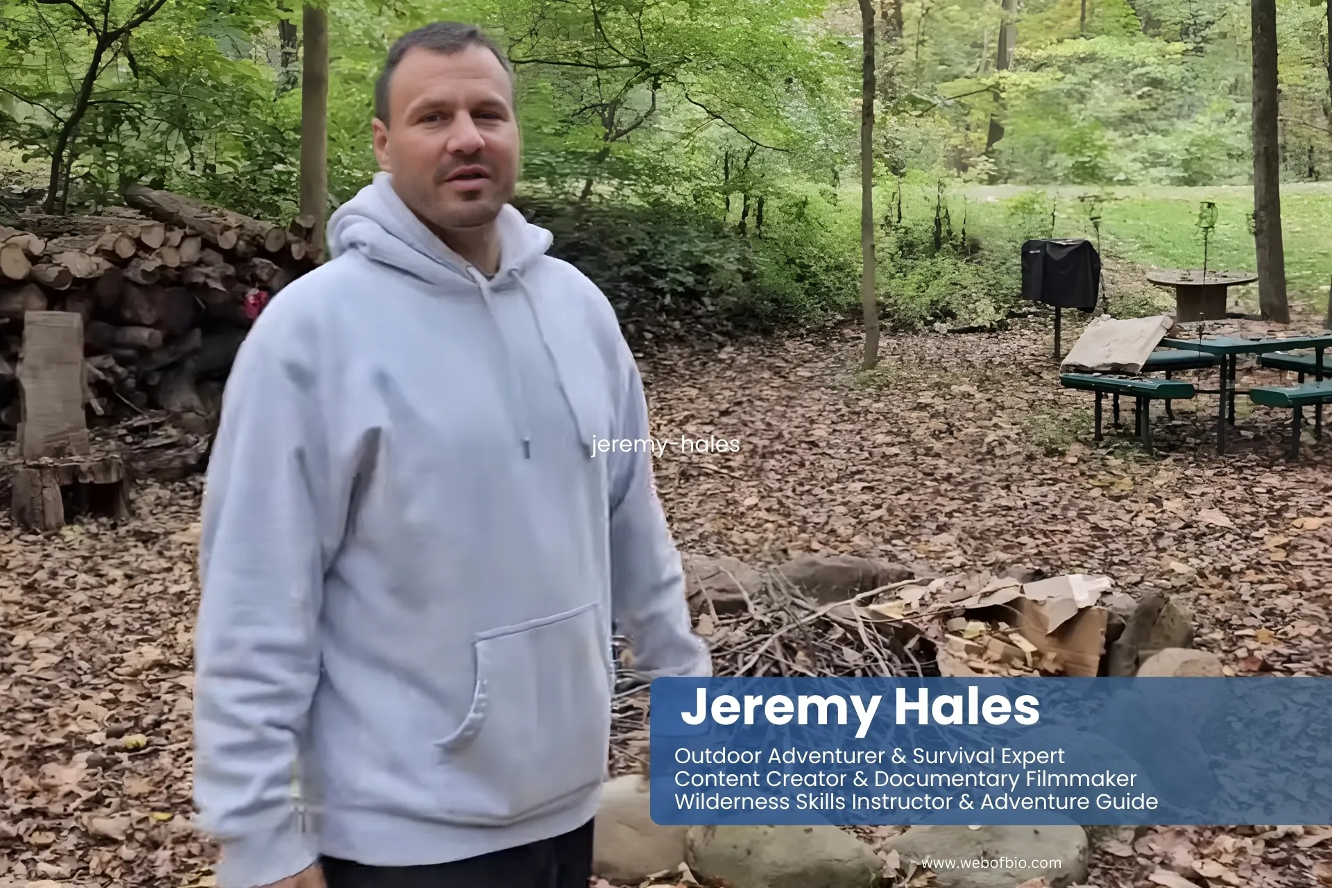 Jeremy Hales stands in a wooded area beside a fire pit, surrounded by trees and natural scenery