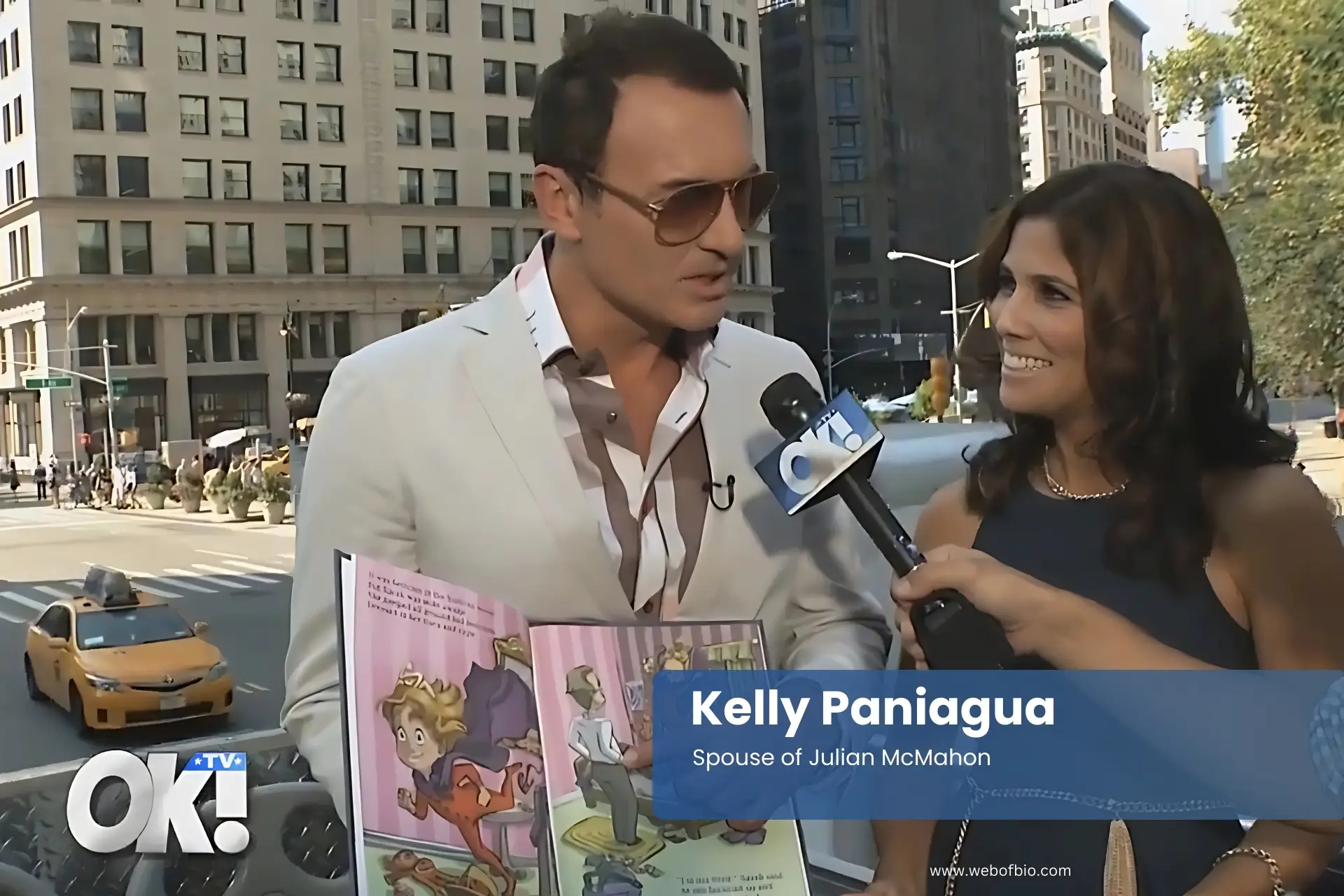 Kelly Paniagua and her husband Julian McMahon in a suit is interviewed by a journalist on the street.