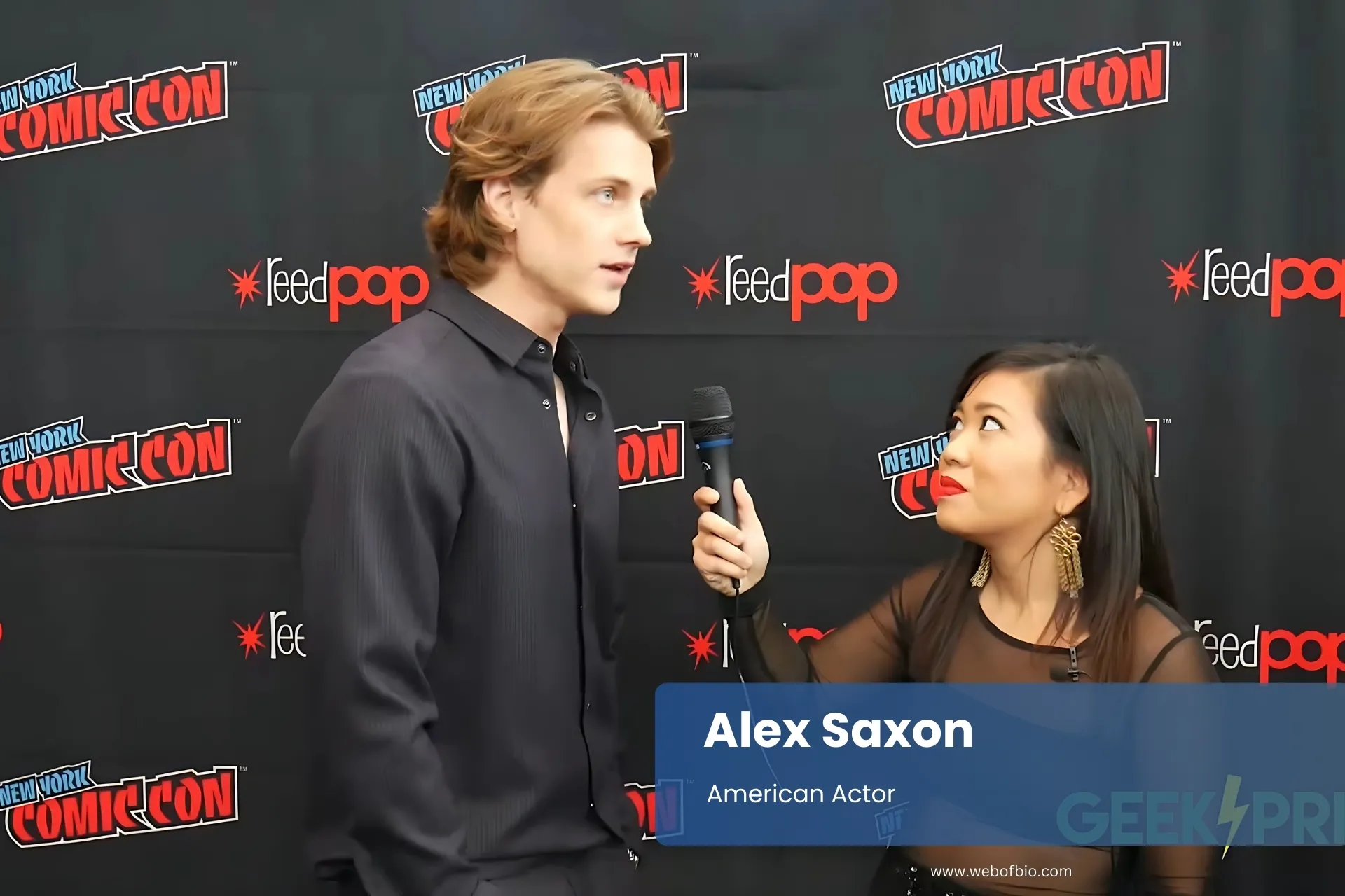 Alex Saxon engage with a reporter in conversation at a comic con, surrounded by colorful comic book displays and enthusiastic attendees.