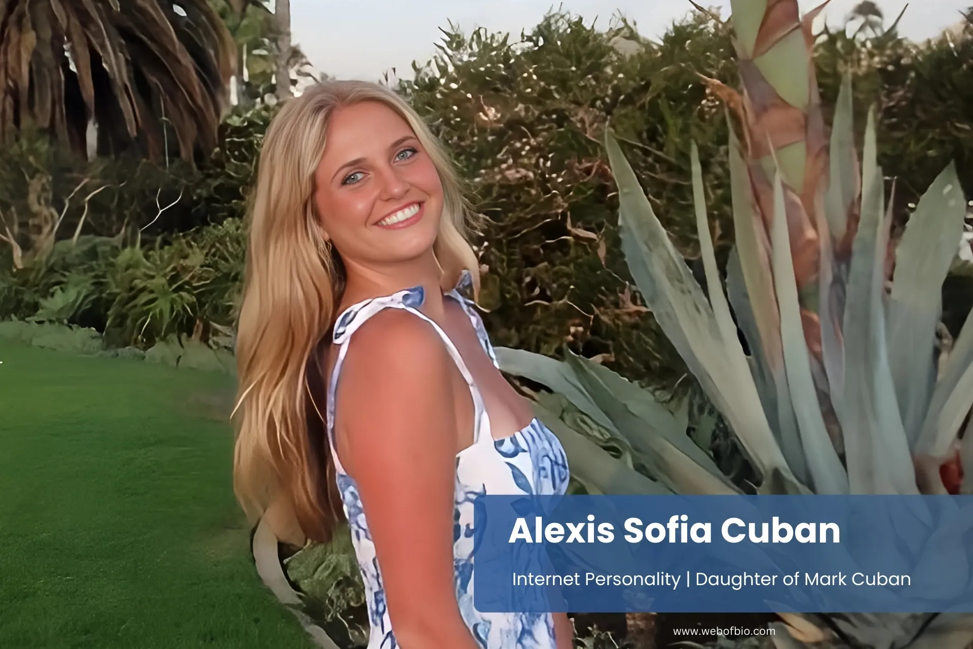 Alexis Sofia Cuban in a blue and white dress stands gracefully in front of a tall palm tree, enjoying a sunny day outdoors.