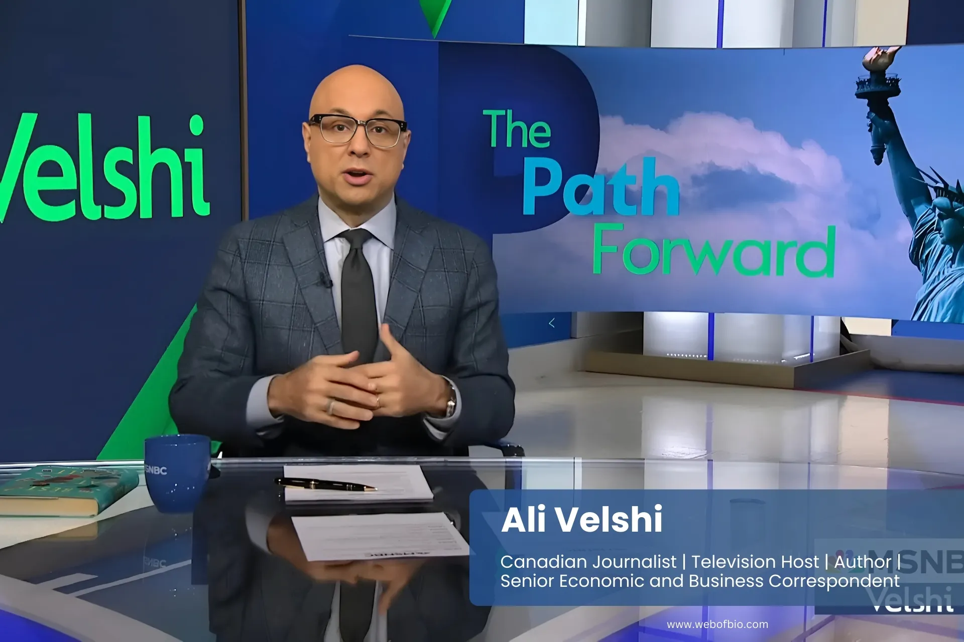 Ali Velshi in a suit and tie sits before a TV displaying the words "The Path Forward," looking contemplative and engaged.