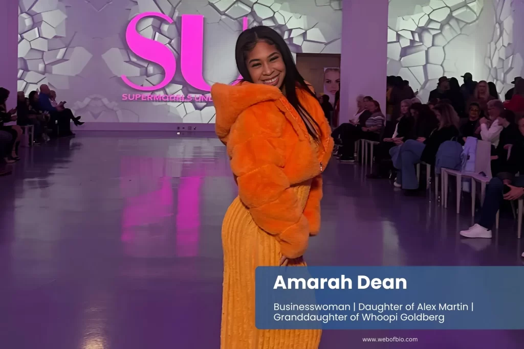 Amarah Dean, wearing an orange fur coat, poses confidently on the runway, showcasing her fashion-forward style.