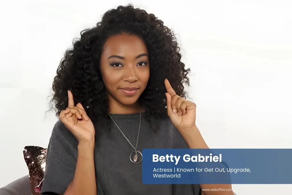 Betty Gabriel sitting on chair with hands raised.