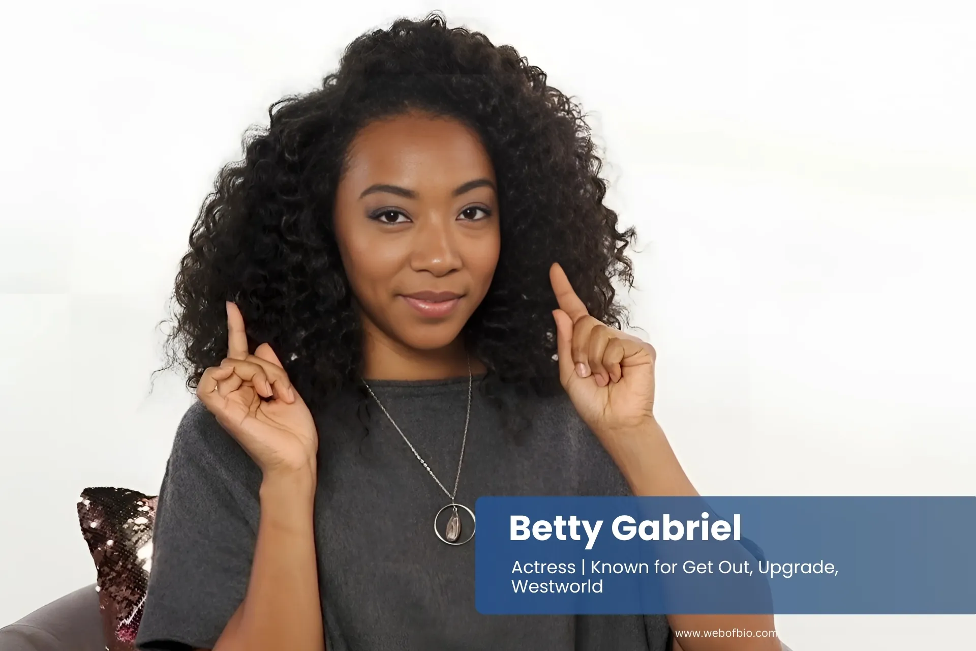 Betty Gabriel sitting on chair with hands raised.