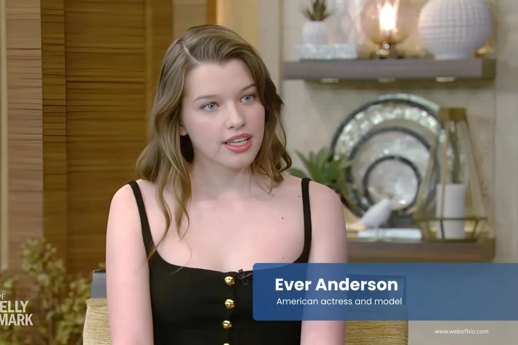 Ever Anderson in a black dress sitting on a chair.