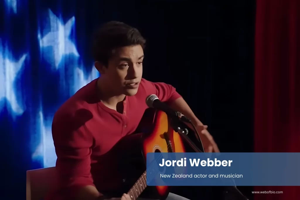 Jordi Webber is playing an acoustic guitar in front of a flag.