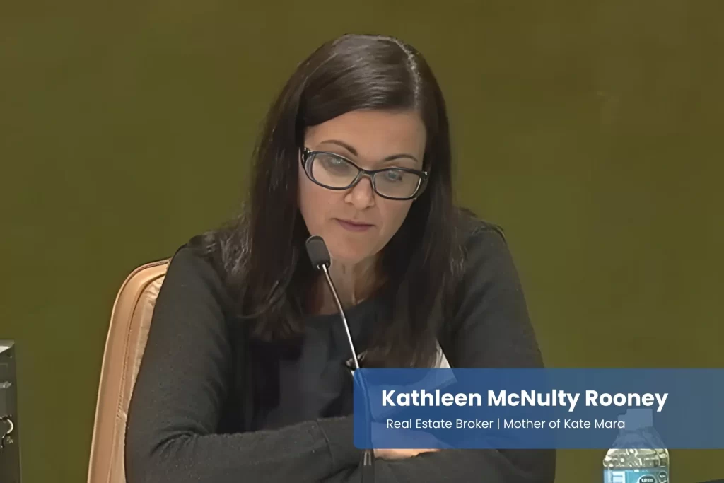 Kathleen McNulty Rooney with glasses sitting at a table in front of a microphone.