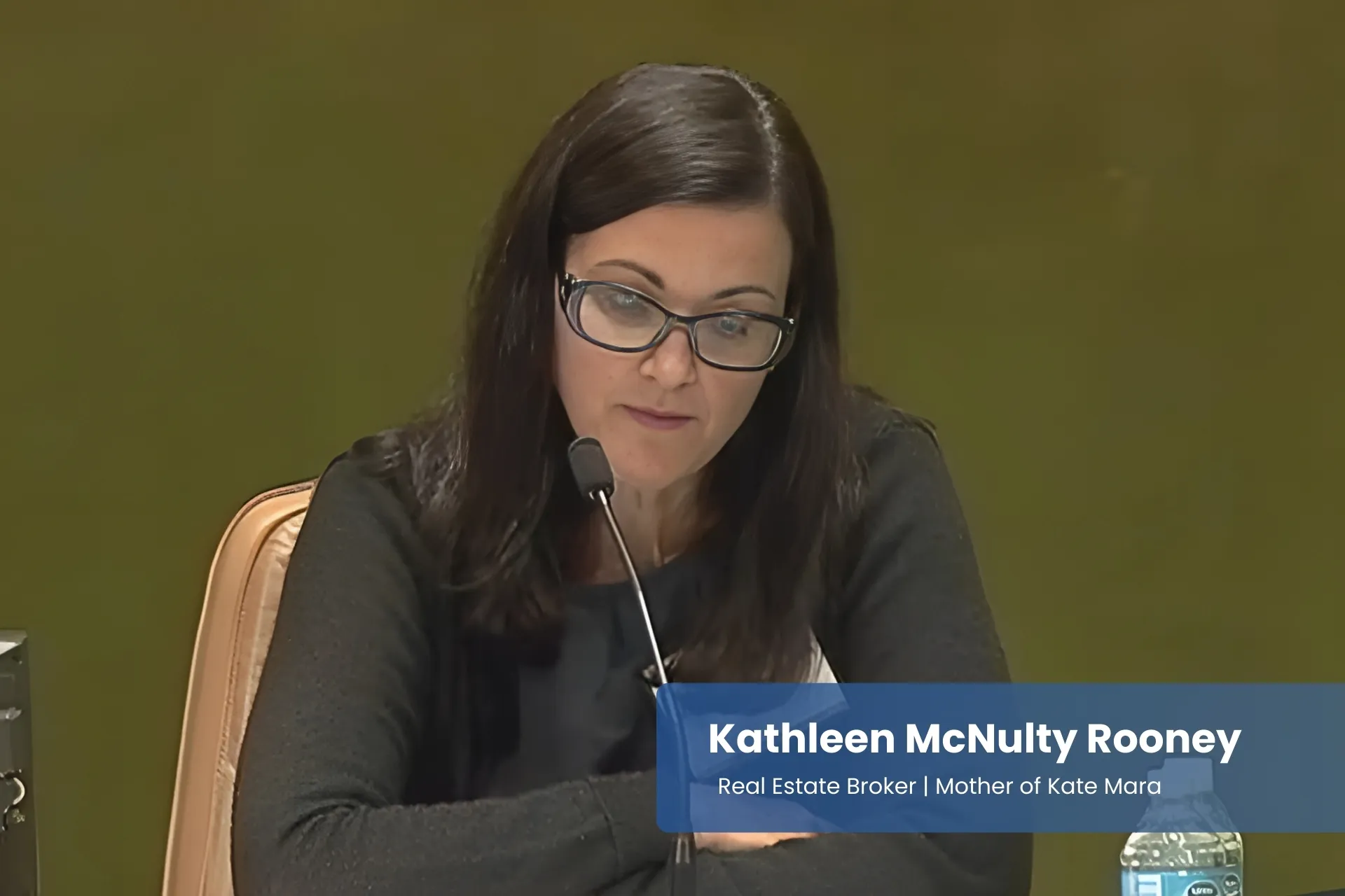 Kathleen McNulty Rooney with glasses sitting at a table in front of a microphone.