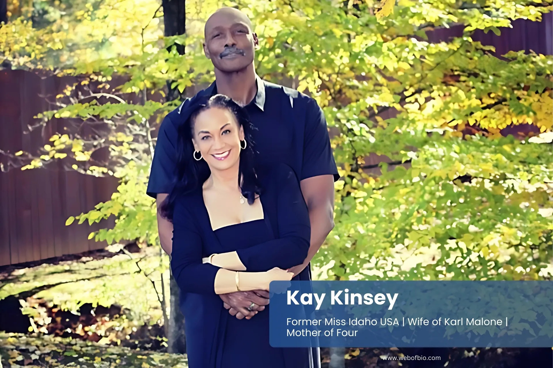 Kay Kinsey and her husband are standing together in a lush green forest, surrounded by tall trees and dappled sunlight.