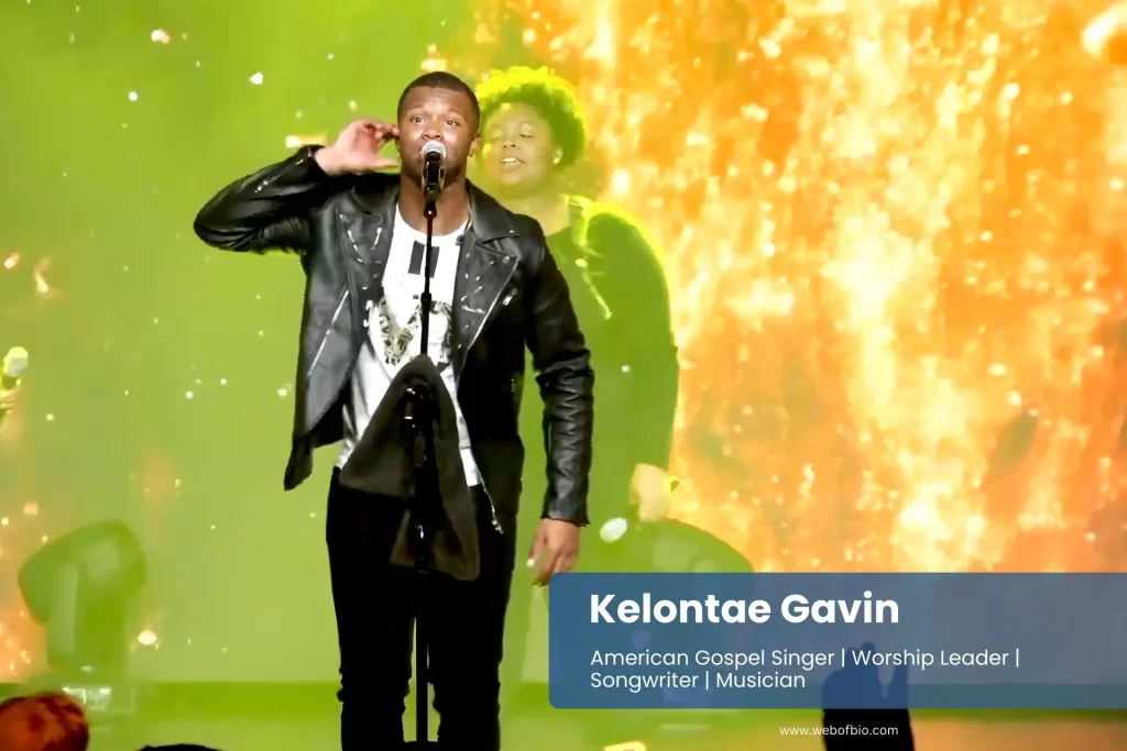 Kelontae Gavin, in a black jacket, stands on stage, addressing a large crowd of people in an auditorium.