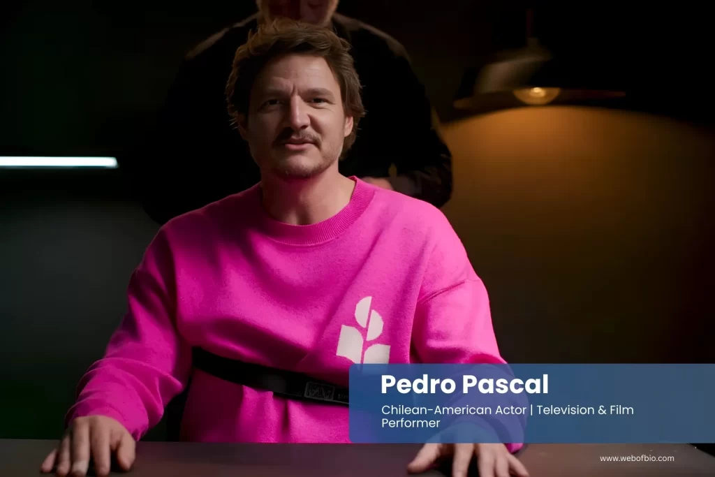 Pedro Pascal in a pink sweatshirt sitting at a table.