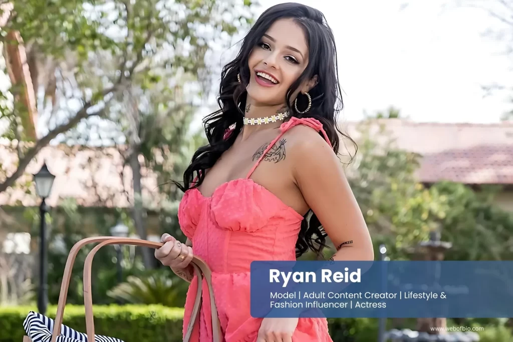 Ryan Reid in a pink dress is holding a handbag.