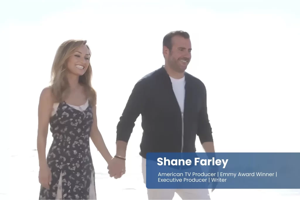 Shane Farley and his girlfriend Giada De Laurentiis stroll hand in hand along a sandy beach, with gentle waves lapping at the shore in the background.