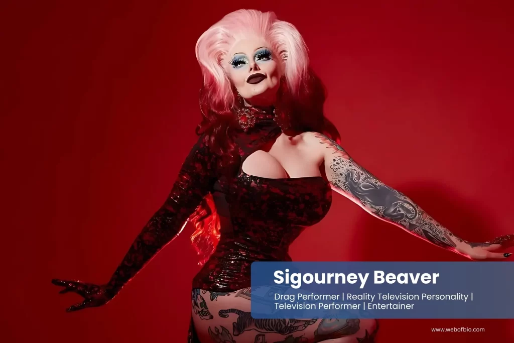 Sigourney Beaver with pink hair and tattoos posing on a red background.