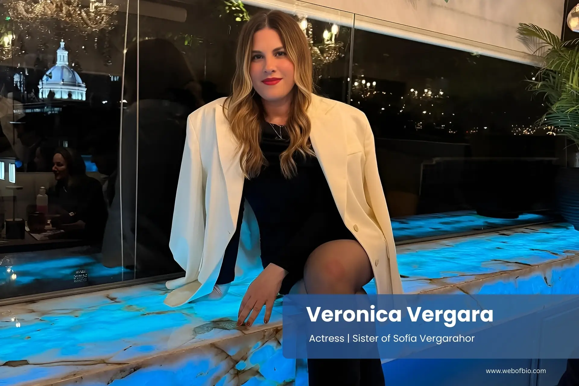Veronica Vergara sitting on a bench in front of a chandelier.